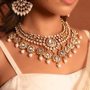 Peut inclure: Un collier doré et une paire de boucles d'oreilles assorties. Le collier présente plusieurs rangées de pierres rondes et transparentes et des pendentifs floraux avec des perles blanches. Les boucles d'oreilles ont un design similaire.