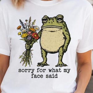 Könnte beinhalten: Weißes T-Shirt mit einer Cartoon-Fröschin, die einen Blumenstrauß hält. Der Frosch ist grün und beige, mit einem mürrischen Gesichtsausdruck. Der Text "sorry for what my face said" ist darunter gedruckt.