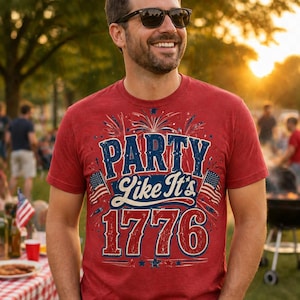 May include: Red t-shirt with the text "PARTY Like It's 1776" in blue and white. The design includes fireworks and American flags. The shirt is worn by a person outdoors, with a blurred background of a gathering.