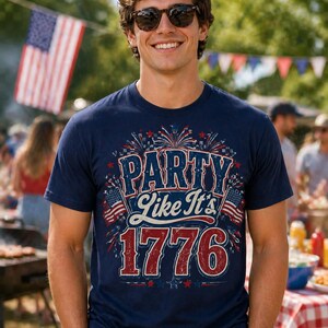 May include: Navy blue t-shirt with the text "PARTY Like It's 1776" in red, white, and blue. The shirt features fireworks and American flags. The person in the image is wearing sunglasses and smiling. An American flag is in the background.