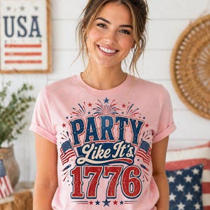 May include: A light pink t-shirt with the text "PARTY Like It's 1776" in blue and red, with fireworks and American flags. The shirt is worn by a person with brown hair.