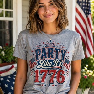 May include: A gray t-shirt with the text "PARTY Like It's 1776" in red, white, and blue. The design includes fireworks, stars, and American flags. The shirt is worn by a person.