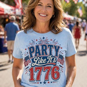 May include: A light blue t-shirt with the text "PARTY Like It's 1776" in red, white, and blue. The shirt features American flags and fireworks graphics, perfect for Independence Day celebrations. The person is smiling in an outdoor setting.