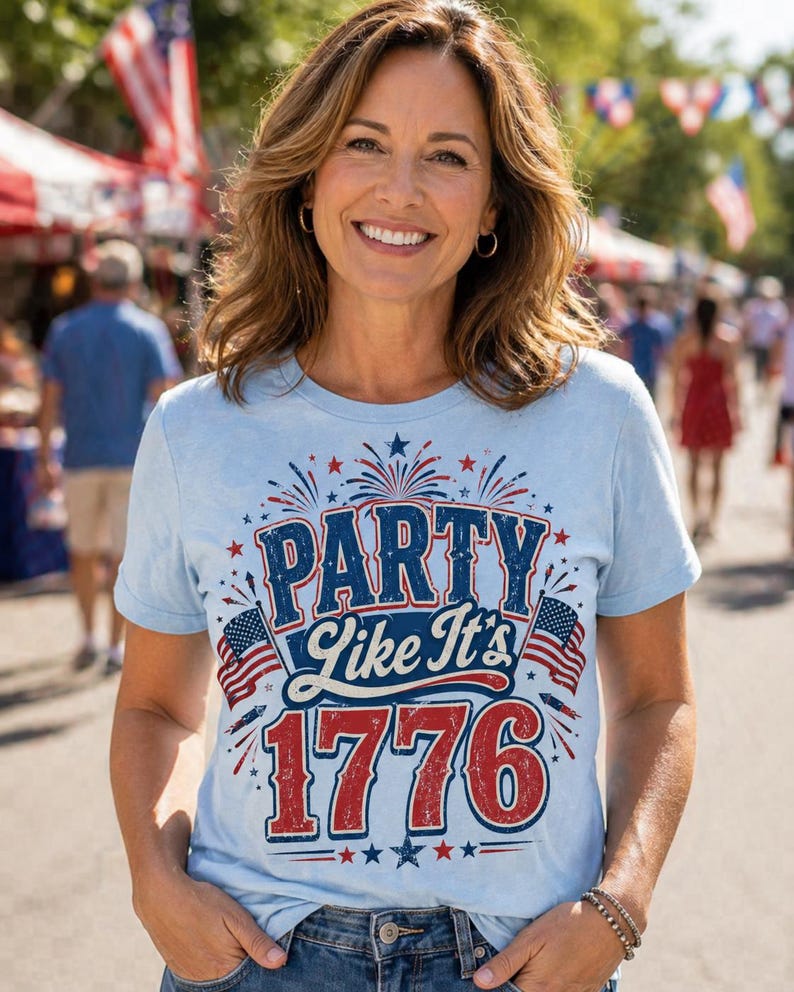 May include: A light blue t-shirt with the text "PARTY Like It's 1776" in red, white, and blue. The shirt features American flags and fireworks graphics, perfect for Independence Day celebrations. The person is smiling in an outdoor setting.
