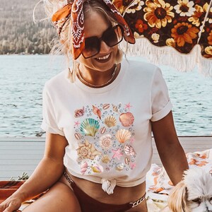 May include: A woman wearing a white t-shirt with a seashell design, a red bandana, and sunglasses sits on a blanket with a small white dog. She is sitting under a floral patterned umbrella.