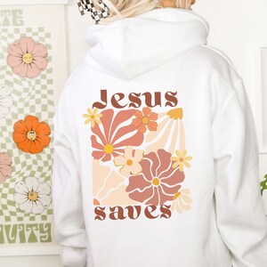 May include: White hoodie featuring a floral design and the words "Jesus saves" in brown lettering. The design includes peach, orange, and yellow flowers. A casual, comfortable garment.
