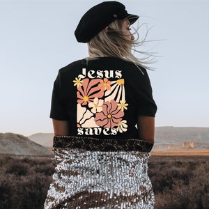 May include: Black t-shirt with a floral design and the words "Jesus saves" in a retro font. The design features flowers in shades of pink, peach, and yellow. The shirt is worn by a person with blonde hair, wearing a black hat.