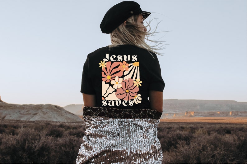 May include: Black t-shirt with a floral design and the words "Jesus saves" in a retro font. The design features flowers in shades of pink, peach, and yellow. The shirt is worn by a person with blonde hair, wearing a black hat.