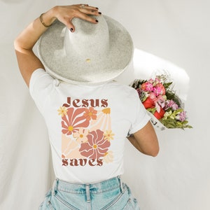 May include: White t-shirt with a floral design and the words "Jesus saves" in a retro font. The design features flowers in shades of orange, peach, and cream. The person is wearing a grey hat and holding a bouquet of flowers.