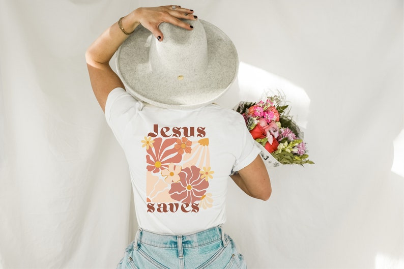 May include: White t-shirt with a floral design and the words "Jesus saves" in a retro font. The design features flowers in shades of orange, peach, and cream. The person is wearing a grey hat and holding a bouquet of flowers.
