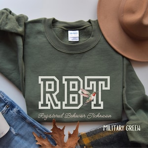 Könnte beinhalten: Ein militärgrüner Pullover mit dem Text "RBT Registered Behavior Technician" und einer Kolibri-Grafik. Der Text ist in einer weißen Blockschrift.