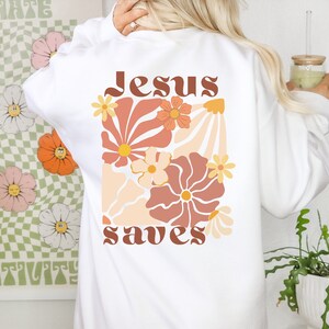 May include: White sweatshirt featuring a floral design in orange, peach, and brown. The words "Jesus saves" are printed in a retro font. The sweatshirt has long sleeves and a crew neck.