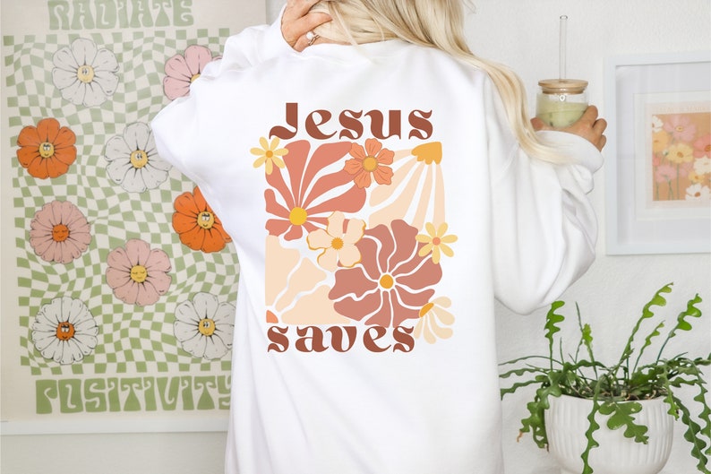 May include: White sweatshirt featuring a floral design in orange, peach, and brown. The words "Jesus saves" are printed in a retro font. The sweatshirt has long sleeves and a crew neck.