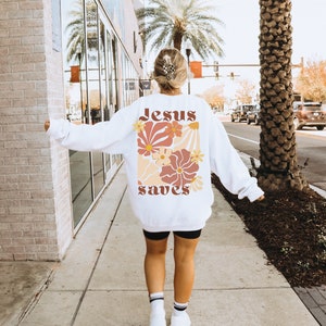 May include: White sweatshirt with a floral design and the words "Jesus saves" in a retro font. The design features flowers in shades of orange, peach, and brown. The sweatshirt is worn by a person walking on a pavement.