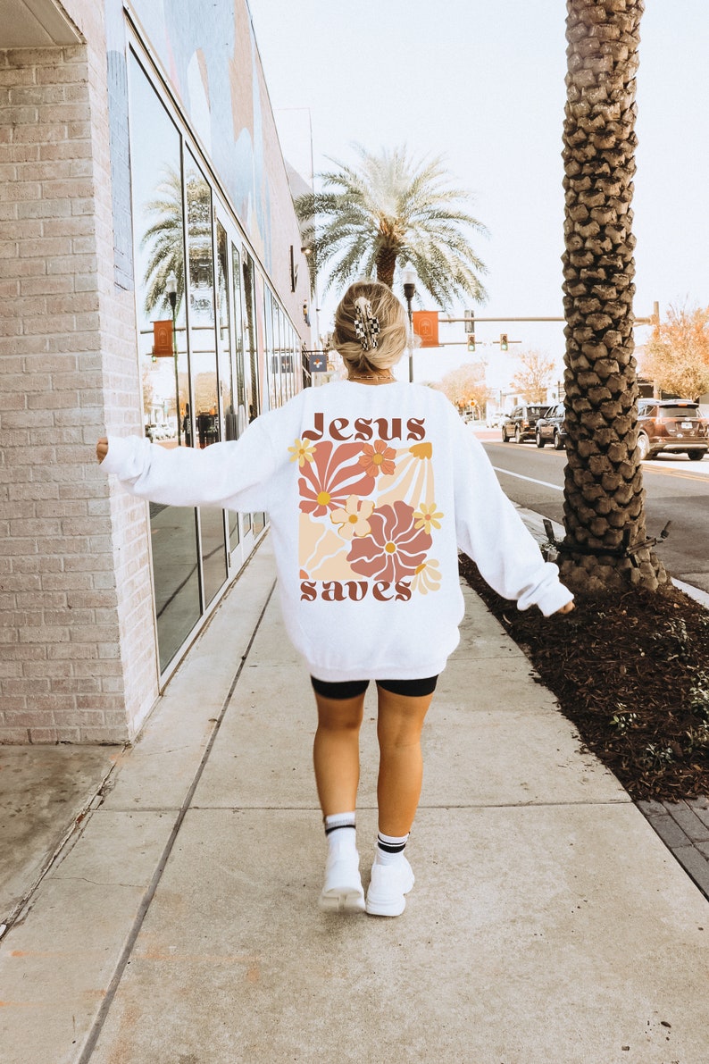 May include: White sweatshirt with a floral design and the words "Jesus saves" in a retro font. The design features flowers in shades of orange, peach, and brown. The sweatshirt is worn by a person walking on a pavement.