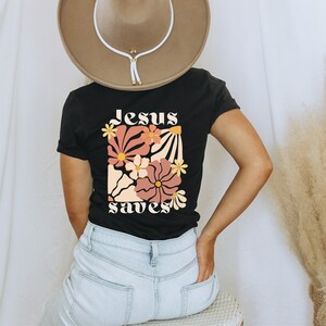 May include: Black t-shirt with a floral design and the words "Jesus saves" in a retro font. The shirt features a colourful flower graphic in shades of pink, orange, and yellow. The person is wearing a brown hat and light blue jeans.
