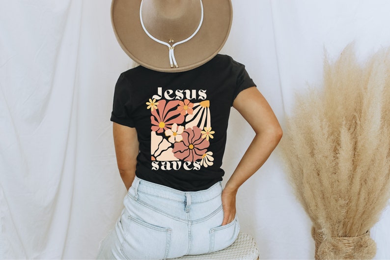 May include: Black t-shirt with a floral design and the words "Jesus saves" in a retro font. The shirt features a colourful flower graphic in shades of pink, orange, and yellow. The person is wearing a brown hat and light blue jeans.