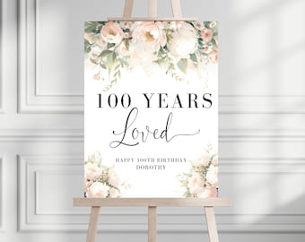 May include: A white sign with a floral design featuring pink and green flowers. The sign reads "100 Years Loved" and "Happy 100th Birthday Dorothy".