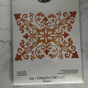 May include: A gold-colored metal die cut with a decorative design. The die cut is shown at 75% of its actual size. The packaging includes the text "Damask #2, Damassé #2, Damasco #2, DIE, POINÇON, TROQUEL, Thinlits™".