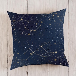 Könnte beinhalten: Quadratisches Kissen mit marineblauem Hintergrund und goldenem Sternbildmuster. Das Kissen zeigt ein himmlisches Design mit Sternen und verbundenen Linien, das einen Nachthimmel-Effekt erzeugt. Auf einer hellen Holzoberfläche platziert.