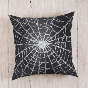 Puede incluir: Cojín decorativo cuadrado negro con un diseño de telaraña blanca. La telaraña es detallada y centrada, con un aspecto texturizado y una zona central más clara. Ideal para decoración de Halloween o gótica.