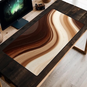 May include: A rectangular desk mat with a swirling design in shades of brown and cream. The pattern resembles flowing waves or layers of paint. The mat is placed on a wooden desk, with a computer monitor and small plants visible in the background.