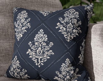 May include: Square decorative pillow with a navy blue background and a white floral pattern. The pillow is placed on a gray sofa, showcasing its design and texture. The floral design is repeated across the pillow's surface.