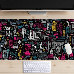 May include: A black rectangular desk mat with a vibrant, abstract design. The pattern features a mix of geometric shapes, lines, and symbols in white, pink, yellow, and teal. A white keyboard and mouse are visible.