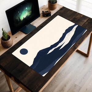 Puede incluir: Una alfombrilla de escritorio blanca con un diseño de montaña azul y blanco. La alfombrilla está sobre un escritorio de madera oscura con un monitor de ordenador y una planta en maceta.