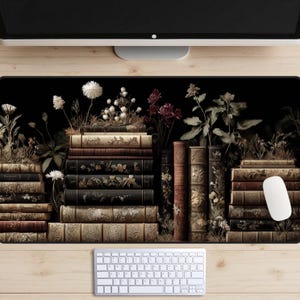 Op de afbeelding: Zwarte bureaumat met een ontwerp van gestapelde antieke boeken en bloemaccenten. De boeken zijn in bruin- en goudtinten, met witte bloemen en groen gebladerte. Een witte muis en toetsenbord zijn zichtbaar.