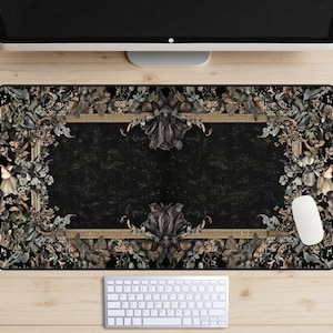Op de afbeelding: Een decoratieve zwarte bureaumat met een bloemen- en sierlijk frame-ontwerp. De mat heeft een donkere achtergrond met een goudkleurig frame en bloemaccenten in tinten groen, grijs en beige. Een computermuis en toetsenbord zijn zichtbaar op een houten bureau.