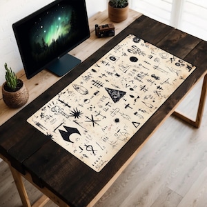 May include: A black and white graphic design desk mat featuring a range of symbols and patterns. The mat is on a wooden desk with a computer monitor and a potted plant.