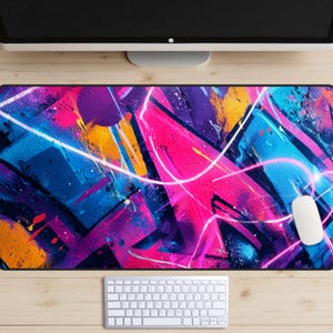 May include: A vibrant mouse pad featuring a graffiti-style design with bold splashes of pink, blue, purple, and yellow. White lines and splatters add detail to the abstract art. A white mouse and keyboard are also visible.