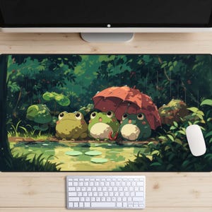 May include: A rectangular mousepad featuring an illustration of three cartoon frogs under a red umbrella in a forest setting. The frogs are green and white, and the background is lush with green foliage and a pond.