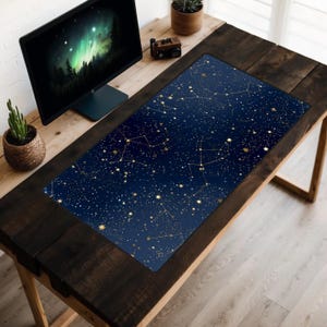 May include: A rectangular desk mat featuring a dark blue background with a gold constellation pattern. The mat is placed on a dark wooden desk, with a computer monitor and small plants visible in the background.