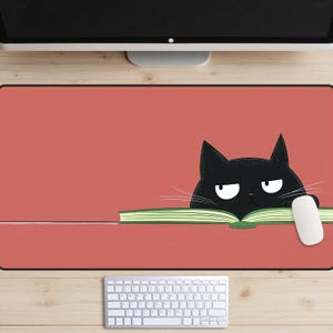 Puede incluir: Una alfombrilla de ratón roja con un gato negro asomándose por encima de un libro verde. El gato tiene una expresión de enfado y mira hacia la derecha. La alfombrilla de ratón está sobre una superficie de madera con un teclado blanco.