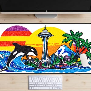 May include: A rectangular mousepad featuring a colorful illustration of Seattle. The design includes a black and white orca leaping from a blue wave, the Space Needle, palm trees, and a sunset. A white mouse and keyboard are visible.