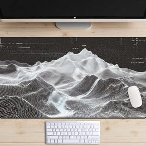 Pode incluir: Um tapete de rato de computador a preto e branco com um design topográfico de cordilheira. Um rato e um teclado de computador brancos são visíveis. O tapete é retangular e está numa secretária de madeira.