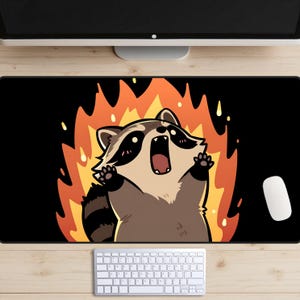 May include: Black rectangular mousepad featuring a cartoon raccoon with a surprised expression, surrounded by orange and yellow flames. The raccoon has brown and tan fur with black markings. A white mouse and keyboard are visible.