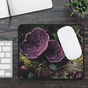 Puede incluir: Alfombrilla de ratón rectangular con una ilustración detallada de setas moradas y follaje forestal. El diseño incluye tonos de verde, marrón y morado, con un borde negro. Un ratón de ordenador blanco descansa sobre la alfombrilla.