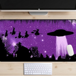 Puede incluir: Alfombrilla de ratón morada con un diseño de Halloween. Muestra siluetas de brujas en escobas, un OVNI, un cielo estrellado, una casa y personas. También se ven un ratón y un teclado de ordenador blancos.