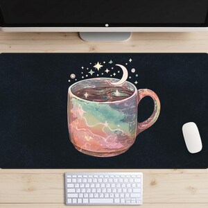 Pode incluir: Tapete de mesa preto com uma ilustração colorida de uma caneca de café com um design celestial. A caneca apresenta um padrão de galáxia, uma lua crescente e estrelas. Um rato e um teclado brancos também são visíveis.