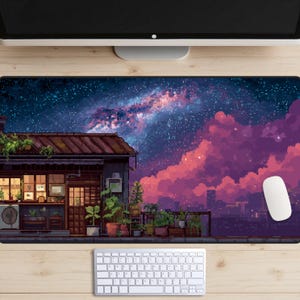 Op de afbeelding: Een pixel art muismat met een nachtelijke hemel met een sterrenstelsel en roze wolken. Een gebouw met een bruin dak en potplanten staat op de voorgrond. Een witte muis en toetsenbord zijn ook zichtbaar.