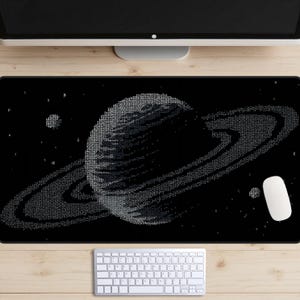 Op de afbeelding: Zwart rechthoekig muismat met een gedetailleerde illustratie van Saturnus en zijn ringen, samengesteld uit kleine, lichtgekleurde symbolen. Een witte muis en toetsenbord zijn zichtbaar onder de monitor. Het ontwerp staat tegen een zwarte achtergrond.