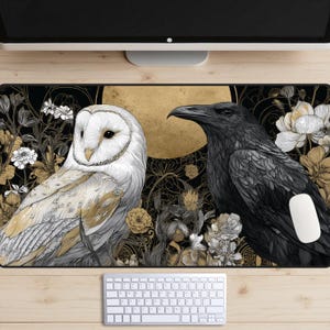 May include: A decorative mouse pad featuring a detailed illustration of a white owl and a black raven, set against a backdrop of a golden moon and floral elements. The design incorporates black, gold, and white colors, creating a striking contrast.