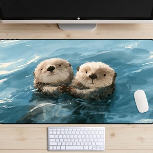May include: A rectangular desk mat featuring a digital illustration of two sea otters floating in blue water, holding hands. The otters have light brown fur, dark noses, and whiskers. The mat is positioned on a wooden desk with a white keyboard and mouse.