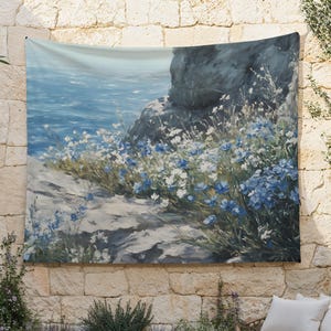 May include: A tapestry depicting a painting of a rocky seascape with blue and white flowers. The flowers are in the foreground, with the ocean and a rocky cliff in the background.