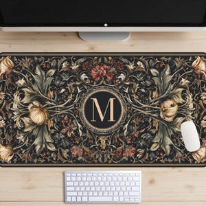 May include: A rectangular mouse pad with a floral design in shades of black, gold, and red. The center features a circular emblem with the letter "M". A white computer mouse and keyboard are visible.