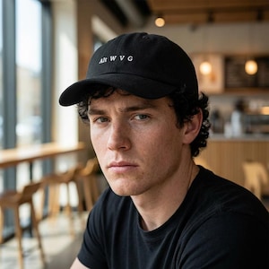 May include: Black baseball cap with the text "Alt WVG" in white. The cap has a curved brim and is worn by a person wearing a black t-shirt. The background shows a cafe setting with wooden chairs and a window.