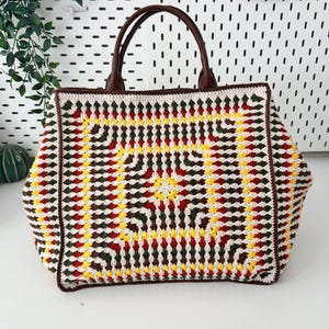 Peut inclure: Un grand sac fourre-tout carré en crochet avec un motif géométrique coloré en rouge, jaune, vert et blanc. Le sac a des poignées et des bordures en cuir marron, et une conception robuste et structurée.